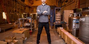 Zachary Hartwig stands arms crossed in what looks like a large workshop. Nitrogen tanks, boxes, ladders, wires, and other materials can be seen in the background.