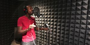 Kelvin Frazier is shown in a recording booth singing into a microphone. The wall shows soundproofing.