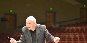 Jonathan Pasternack is shown conducting. He holds a baton and the seats of a theater can be seen in the background.