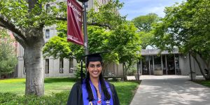 Tara Venkatadri appears in her graduation cap and gown in front of McCormick Hall at MIT. A red banner with the dorm name is attached to a light pole behind her.