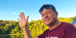 Headshot of Andy Obst ’90 waving while on a boat, sunglasses perched on his head. He's wearing an MIT t-shirt