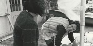 A sailor in a life preserver bends over a boat while looking up at another person whose back is to the camera. A sail flaps in the background.