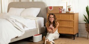 Loewen Cavill sits on the floor next to a bed holding a small device. There is a larger device on the floor next to her and a pad on the bed. Behind Cavill, a cardboard box rests on a chest of drawers.