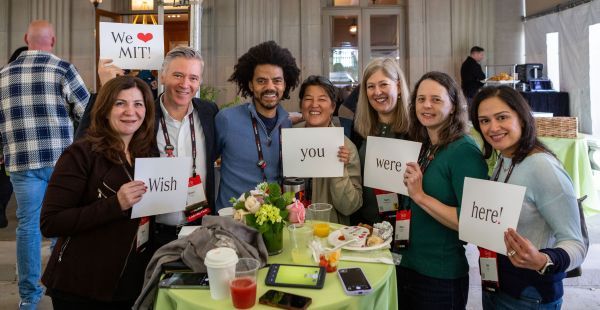 Image: Group of seven alumni standing around a table holding signs that read Wish You Were Here! We Heart MIT!