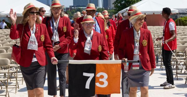 Image: Alumni from the class of 1973 processing into Commencement wearing Cardinal & Gray red coats holding a '73 class banner.