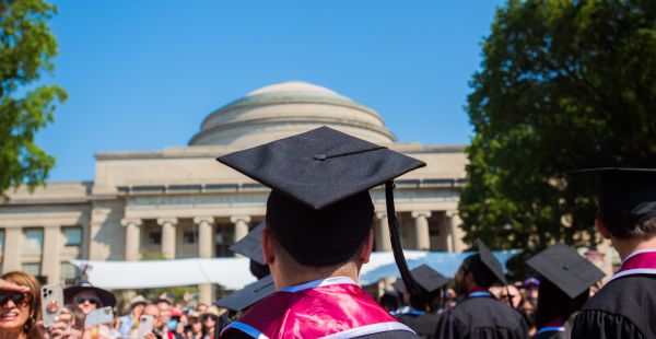 Graduate standing in front of MIT Dome facing away from the camera wearing a graduation robe and cap