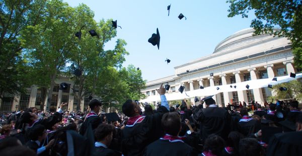 Crowd of graduates in Killian Court cheering and tossing graduation cap with MIT Dome in background.
