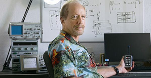 Charlie Holdaway is shown from elbows up, sitting at a desk and holding a keypad that has an antenna. Behind him on the left, machinery and a circular lamp can be seen. On the right, there is a computer screen; Behind him, there is a whiteboard with some numbers and figures visible.