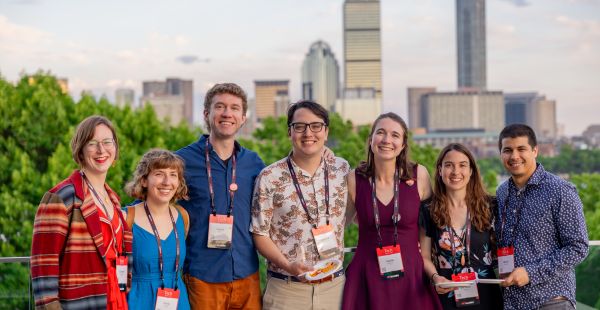 A group of seven smiling individuals poses outdoors with a city skyline in the background, wearing Tech Reunions badges.
