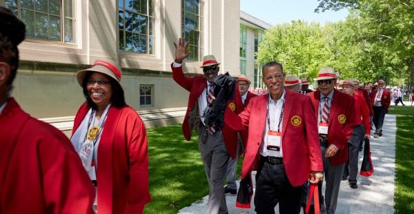 A group of people in red blazers and hats cheerfully walk together outdoors, waving and smiling in front of a building.