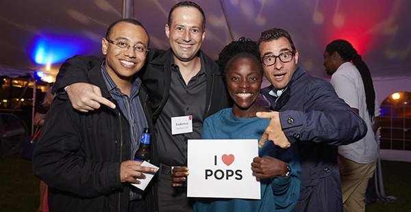 Four alumni standing in a group holding a sign reading "I heart Pops"