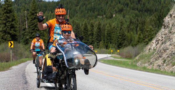 Photo of Steve Zuckerman (waving) and Deb Meyerson riding a tandem bike on a road with trees in the background and another biker behind them