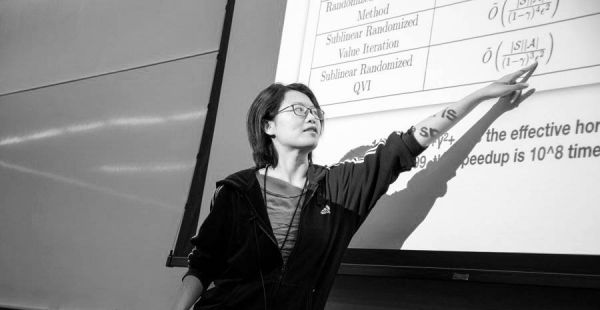 Photo in black and white of Mengdi Wang pointing with her left arm up at a projection screen with equations on the board
