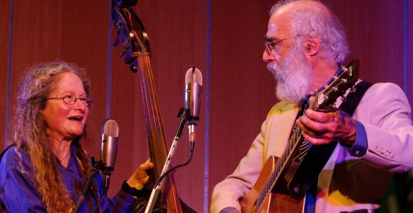 Sharon Horovitch, left, and Jim Muller are shown from the waist up with microphones in foreground. Horovitch is holding the neck of a bass fiddle. Muller is playing a guitar. They are looking at each other.
