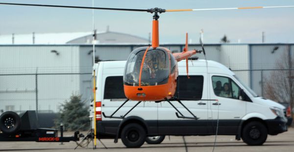 A small orange helicopter hovers in the air just a few feet off the ground. A large white van can be seen behind it with an attached trailer. Two people can be seen looking out the van's window. Beyond the van is a chain-link fence and further still are some industrial buildings.
