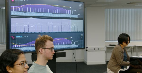A large screen showing a variety of charts is at left in a room where Elaine Chew is playing piano and two students, at left, listen.