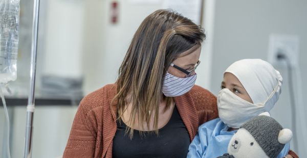 A mother hugs her young daughter while she receives cancer treatment. Both are masked, and the child holds a plush toy