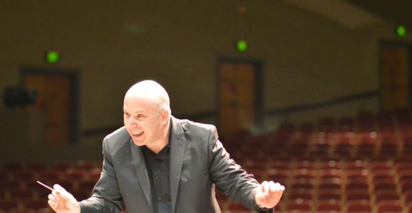 Jonathan Pasternack is shown conducting. He holds a baton and the seats of a theater can be seen in the background.
