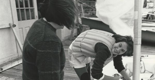 A sailor in a life preserver bends over a boat while looking up at another person whose back is to the camera. A sail flaps in the background.