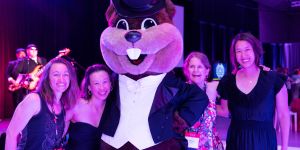Four women in evening dress pose with Tim the Beaver dressed in a tux. Lighting is low and a band member can be seen in the background.