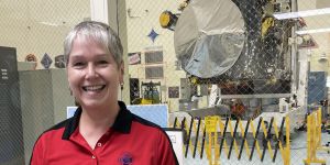 Lindy Elkins-Tanton poses in a red, collared Psyche shirt. A large silver disc on the spacecraft can be seen through the window behind her.