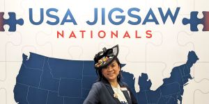 A photo of Tammy McLeod standing in front of a backdrop that says USA Jigsaw Nationals with a map of the US under the words